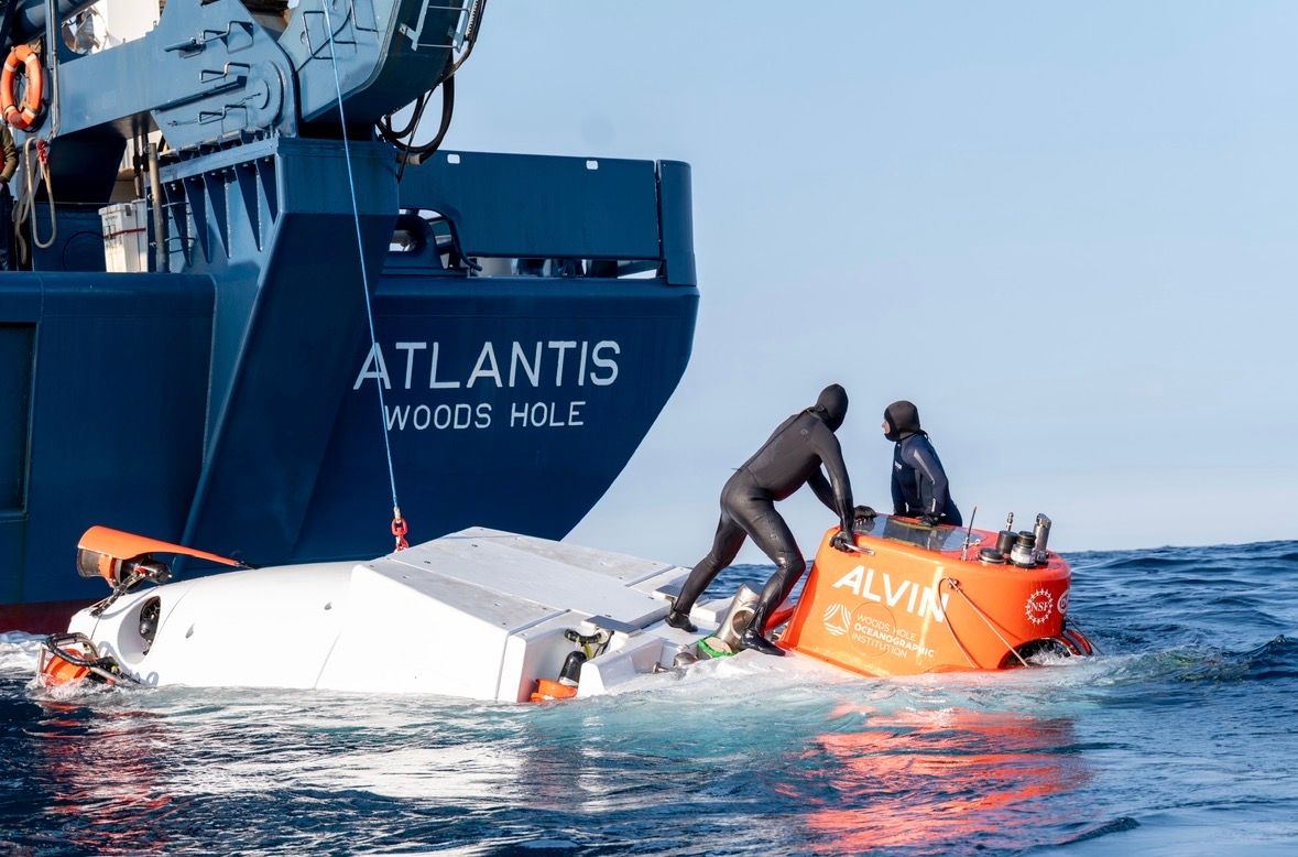 A picture of the submersible Alvin in the water off the back of the Atlantis. Two people in black wetsuits are standing on top of the sub.
