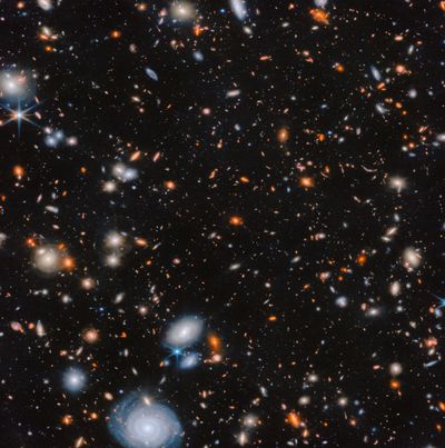 An image of many glowing galaxies in deep space, in various shapes and colors, on a black background. There are some large, blue spiral galaxies, some large and pale white elliptical galaxies, and many orange and red, medium-sized galaxies. Even smaller galaxies, down to tiny faint spots, appear in all these colors.