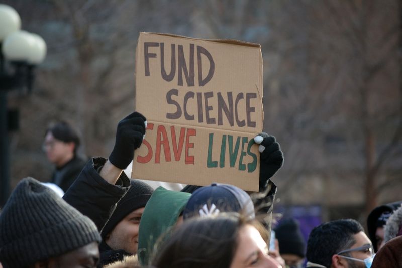 A photo of the January 2025 protests in New York against science funding cuts by Donald Trump's government. One person holds a sign that says "Fund Science, Save Lives".