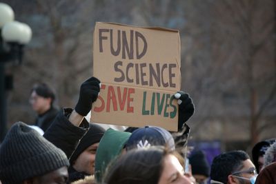 A photo of the January 2025 protests in New York against science funding cuts by Donald Trump's government. One person holds a sign that says "Fund Science, Save Lives".