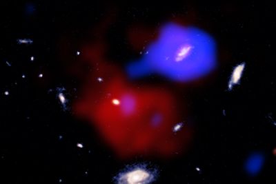 several galaxies are visible in these image. the central two are surrounded by two clouds, one large and red and the other cool and blue. The blue one extends towards the red one with a jet