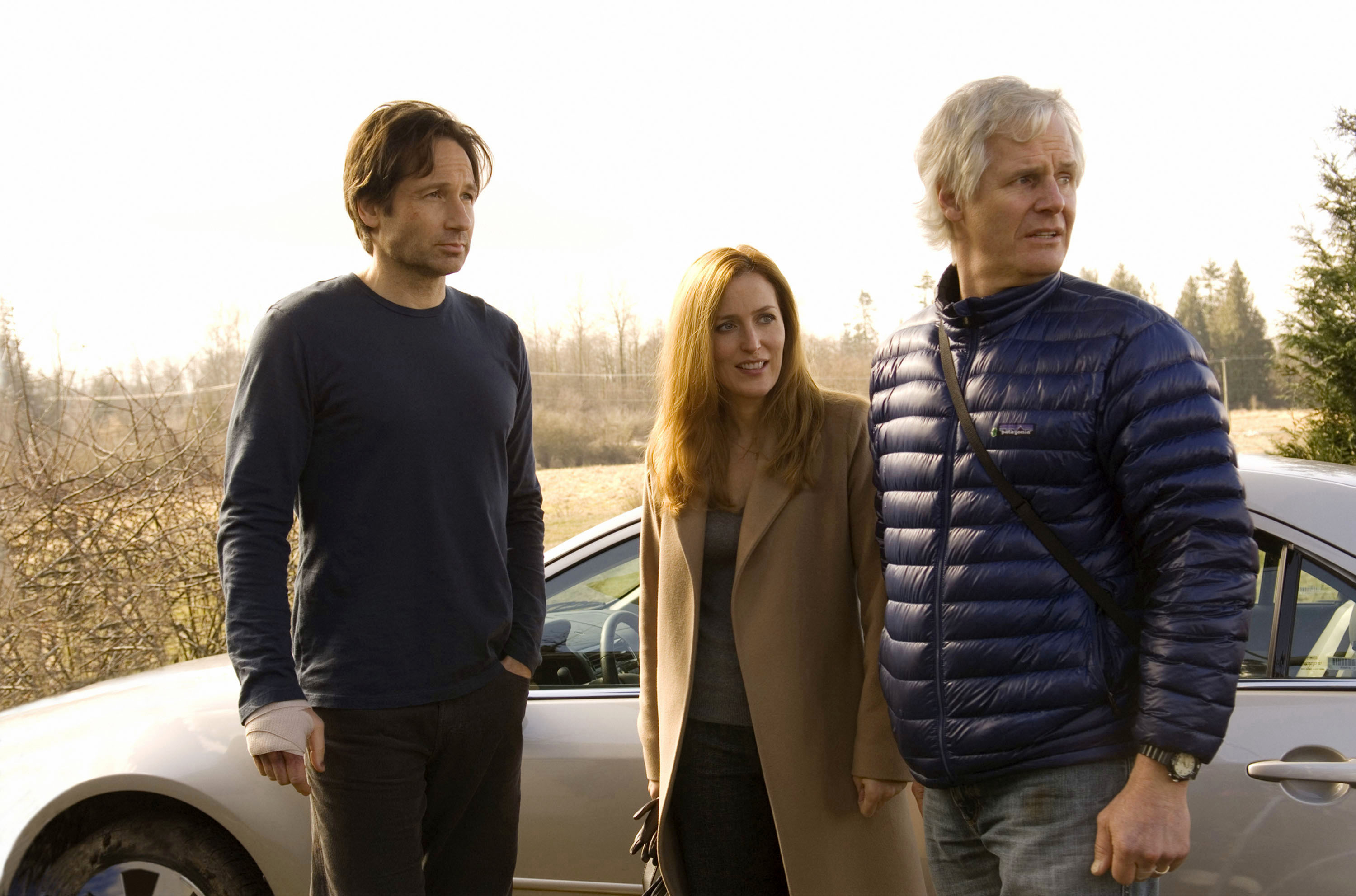 The three of them are looking at something off camera hile standing next to a car in a field The three of them are looking at something off camera hile standing next to a car in a field