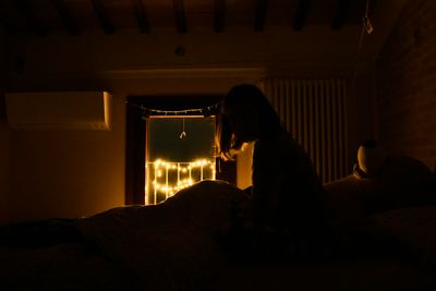woman sitting up in bed in a dark room with some yellow fairy lights in the window