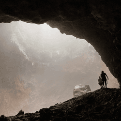 Two silhouetted explorers standing inside massive cave chamber with misty light. The powerful contrast between the pitch-black foreground and the glowing atmospheric haze creates a deeply cinematic and emotionally compelling scene. A striking visual metaphor for human insignificance against nature, discovery, adventure, and the unknown. Ideal for editorial use, travel campaigns, adventure brands, book covers, and inspirational content.