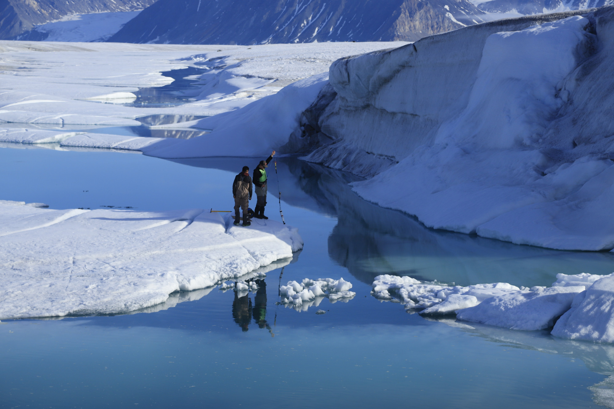 Researchers Joseph Shoapik and Jérémie Bonneau lower instruments through cracks in the ice shelf to measure temperature and salinity at different depths in the water below. Researchers Joseph Shoapik and Jérémie Bonneau lower instruments through cracks in the ice shelf to measure temperature and salinity at different depths in the water below.