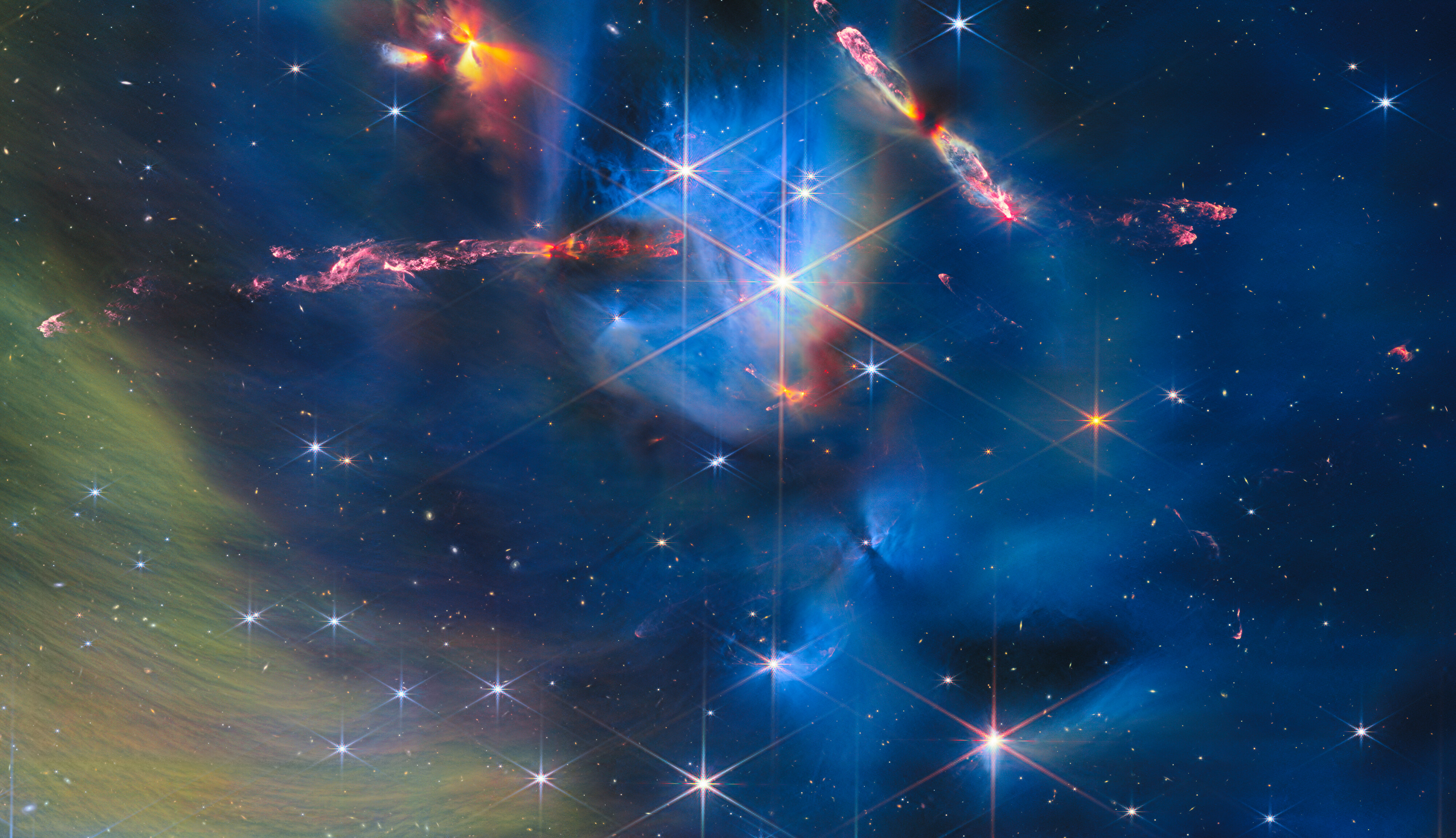 A star-forming nebula that contains young stars called protostars. Two of these protostars are shooting out long jets that are colliding with the nebula, creating glowing, colourful outflows. A few other protostars create orange and red areas in the nebula. Most of the image is covered by blue gas, with brighter areas, denser clouds and dark gaps; the bottom-left corner contains greenish-yellow gas.