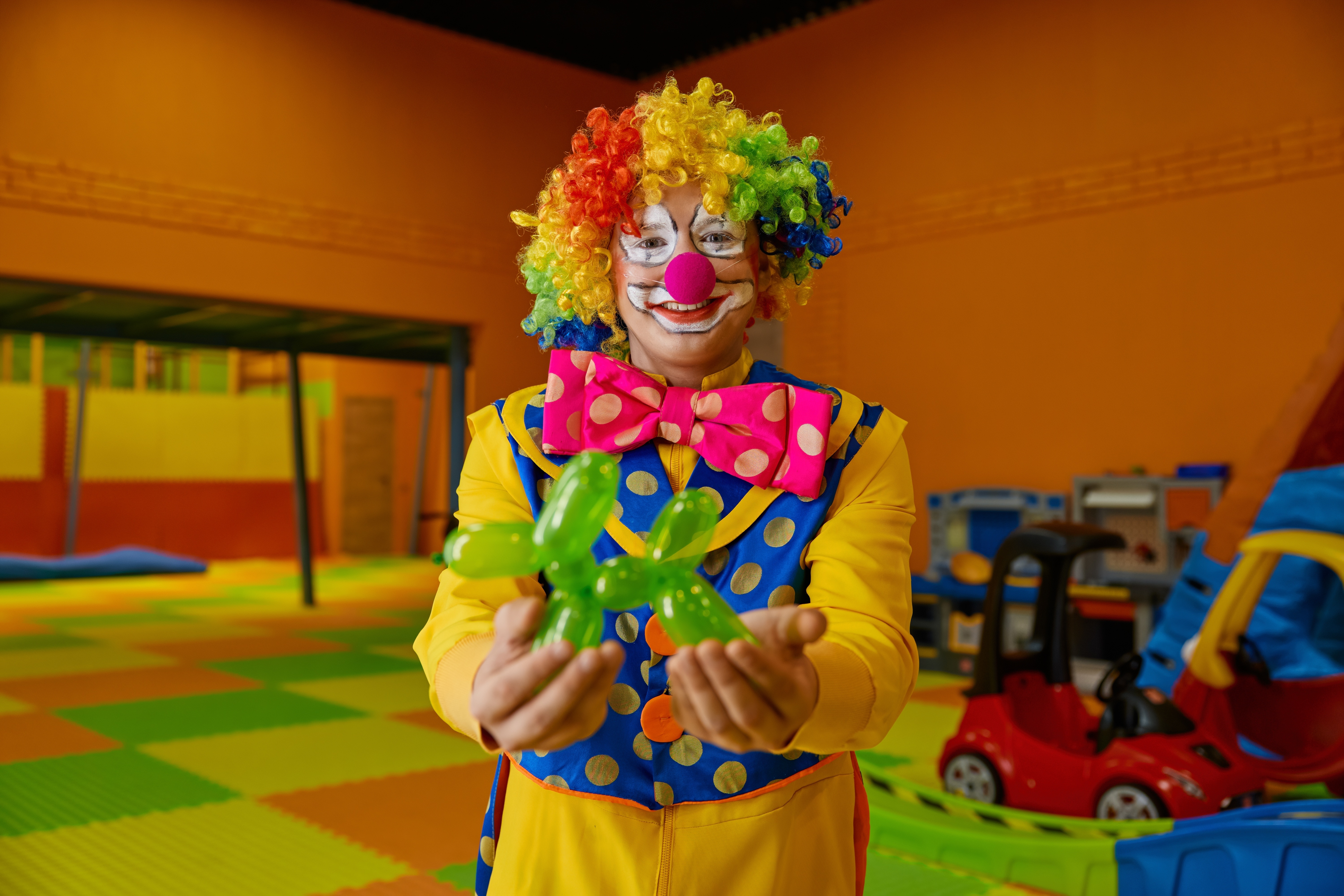 A man dressed as a clown in brightly coloured clothes holding a balloon dog in his hands. A man dressed as a clown in brightly coloured clothes holding a balloon dog in his hands.