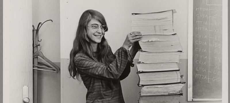 Cropped sepia image of a young Margaret Hamilton, a white woman with long hair and glasses, standing next to a large stack of books as tall as her