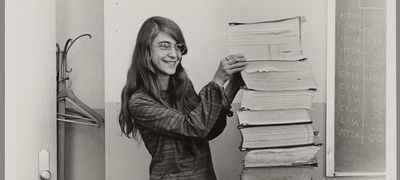 Cropped sepia image of a young Margaret Hamilton, a white woman with long hair and glasses, standing next to a large stack of books as tall as her