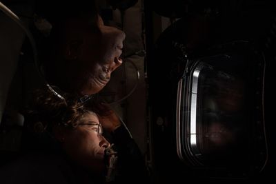 The tip of the heads of the two astronauts are touching as they look out of the window