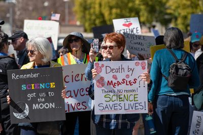 Demonstrators at a pro-science march, one is holding a black sign that reads "REMEMBER, SCIENCE GOT US TO THE MOON" and the the other is holding a white sign that reads "DEATH BY MEASLES? THERE'S NO REASON! SCIENCE SAVES LIVES!"