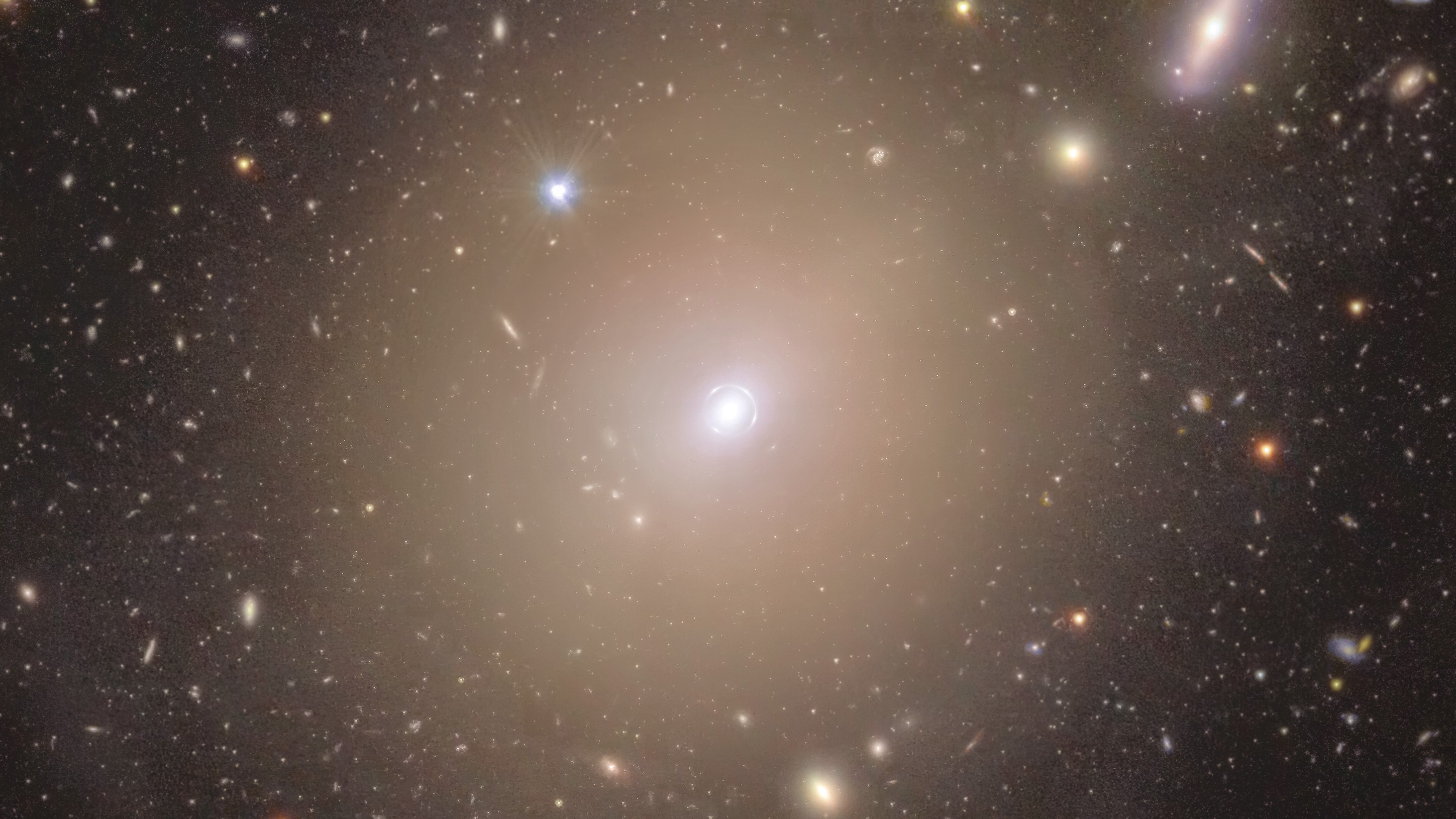 A sea of colourful stars and galaxies appear to swim in the vast blackness of space around a hazy halo at centre stage. In the middle of the image, the fuzzy-looking bulb of light in a warm shade of yellow extends around a small bright spot, nestled within a thin light circle that appears to be drawn closely around it. As we follow the central halo’s rim outwards, its brightness dims and blends smoothly into its surroundings. Here, extended discs of shades ranging from a warm purple to golden yellow, and piercing dots of light with sharp diffraction spikes are spread evenly across the image