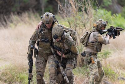 one soldier supports an injured soldier while a third stands behind them aiming his firearm away from them